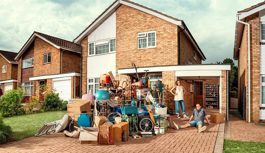 A house with a large amount of clutter and furniture in the front yard, with a man and woman sitting on the porch, surrounded by boxes and other items.