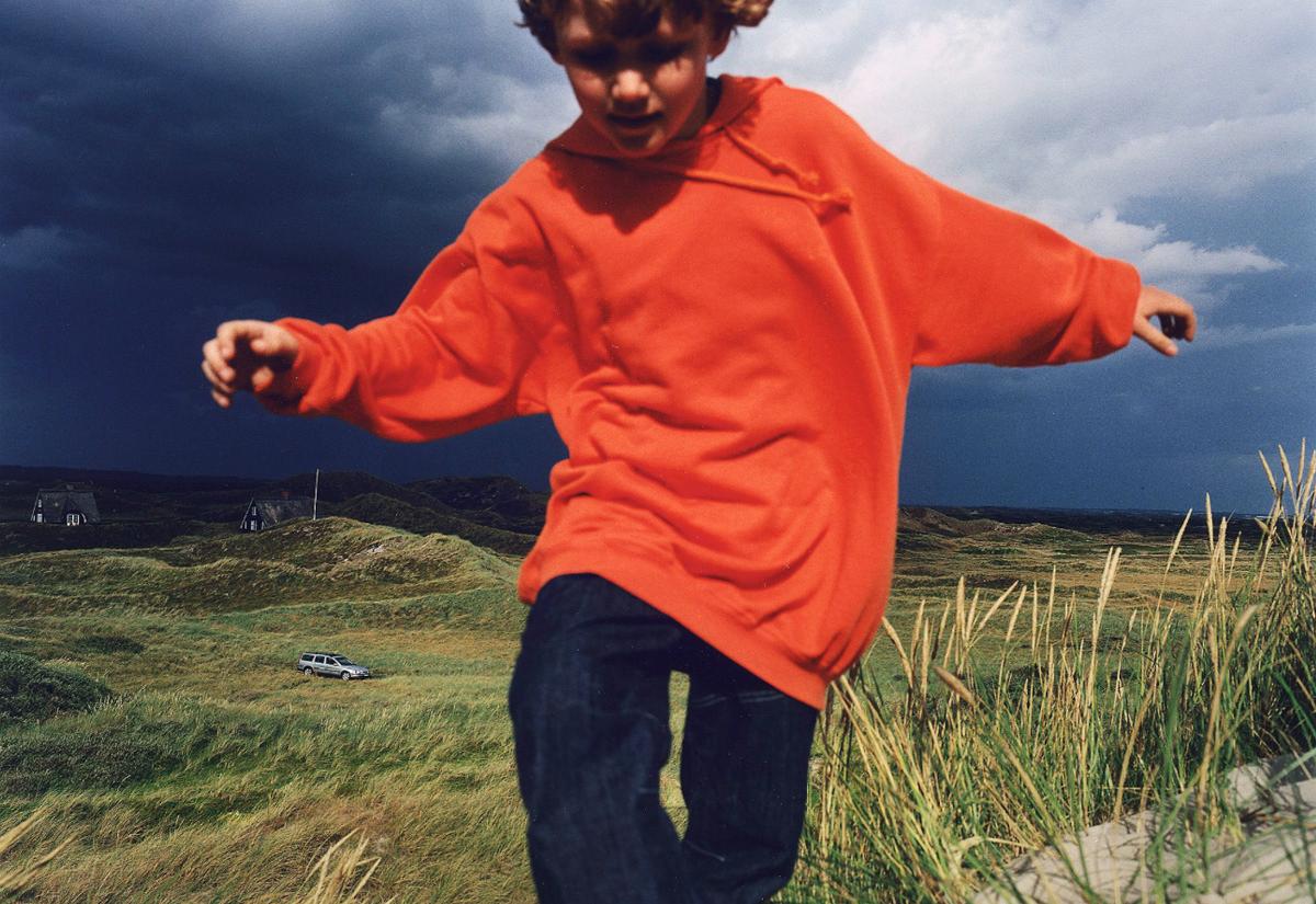 A young boy in an orange hoodie and jeans is running in a field, with a house and a car in the background.