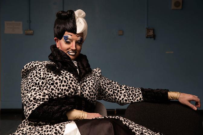The Ugly Sisters, a drag duo, are dressed in matching leopard-print coats with ruffled collars and matching wigs, posing in a dimly lit, industrial-style room.