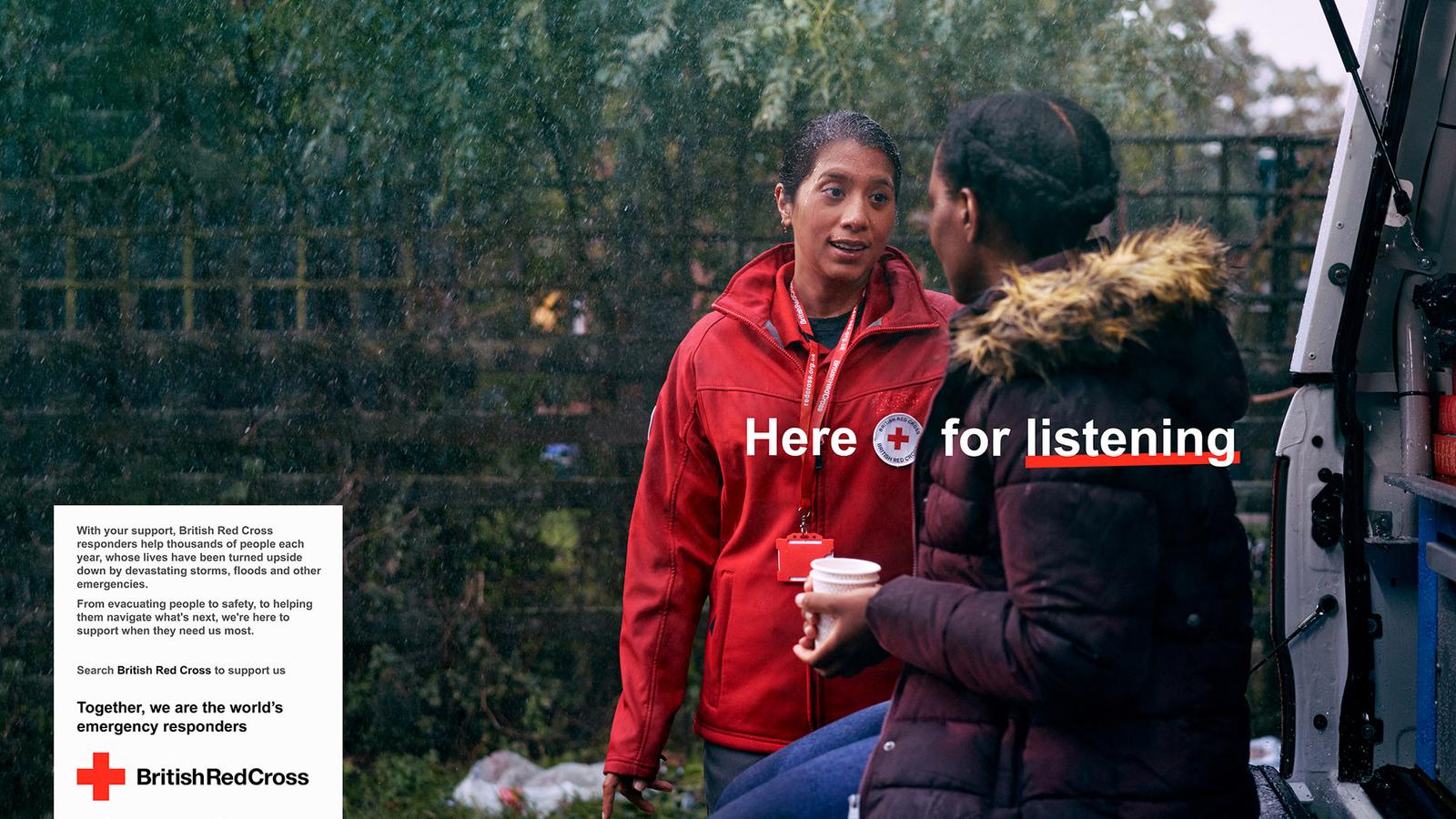 A woman in a red British Red Cross jacket is talking to a woman in a maroon puffer jacket, both standing in front of a van in a rainy, green, outdoor setting.