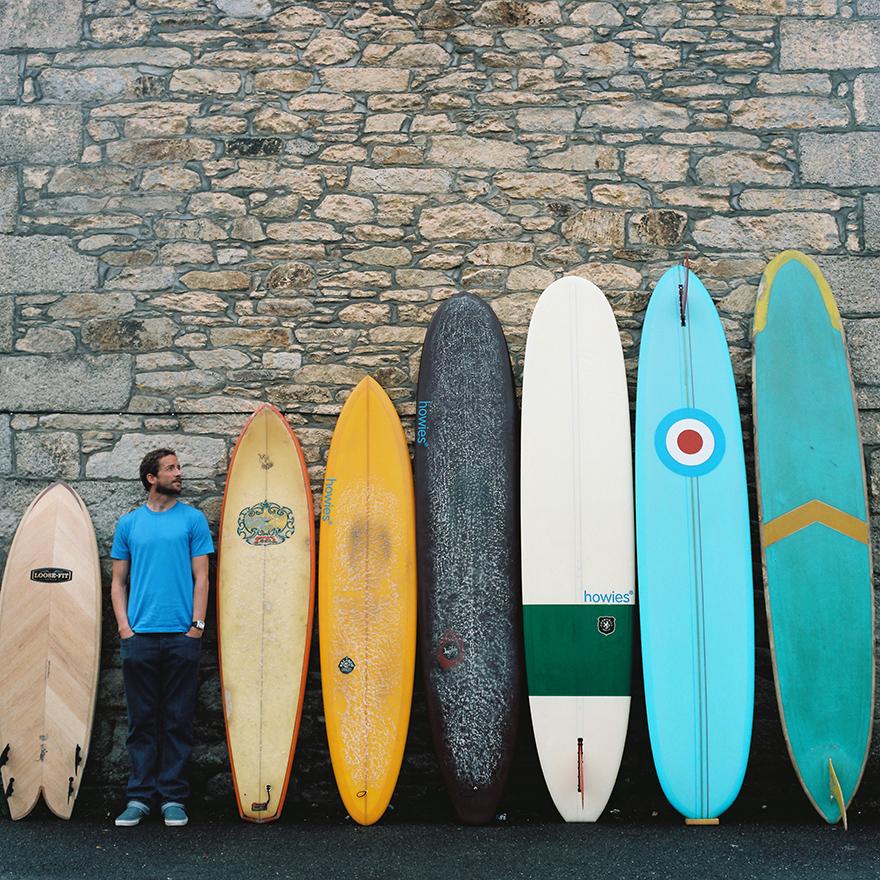 A man in a blue t-shirt and jeans standing next to a wall of surfboards.