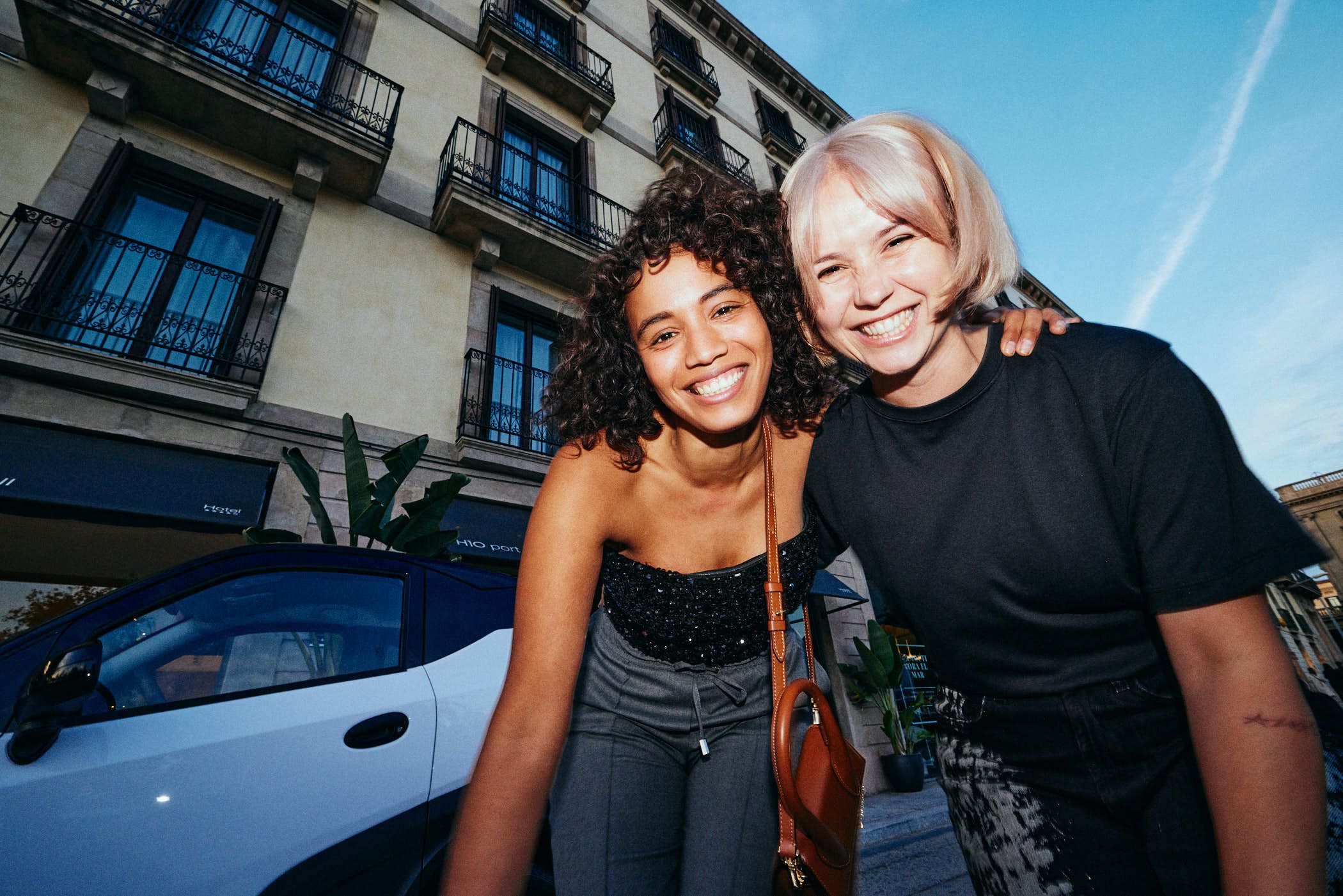 Two women smile at the camera in front of a cream-colored building with black ironwork balconies. The woman on the right has her arm around the woman on the left.
