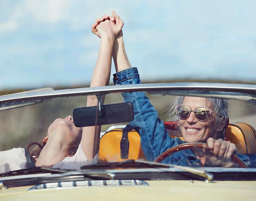 A man and woman in a convertible car raise their arms, smiling.