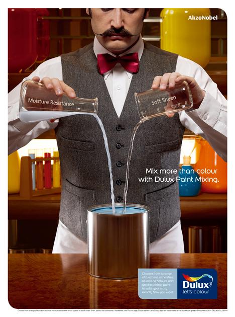A man in a white shirt, black waistcoat, and red bow tie is pouring paint from two bottles into a large metal container.