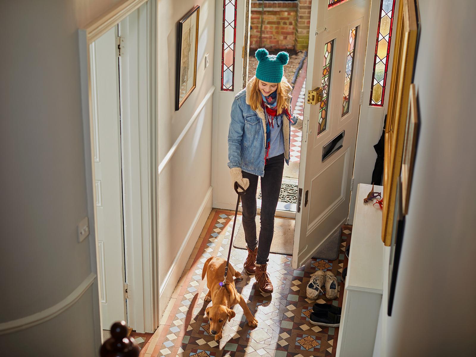 A woman in a blue denim jacket, black trousers, and brown boots walking a small brown dog in a hallway with a patterned floor.