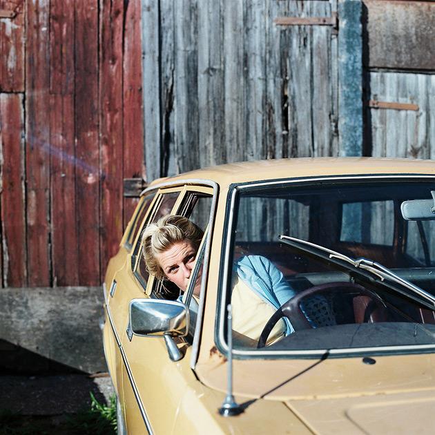 A woman with blonde hair and a blue jacket is sitting in the driver's seat of a yellow car, looking out the window.