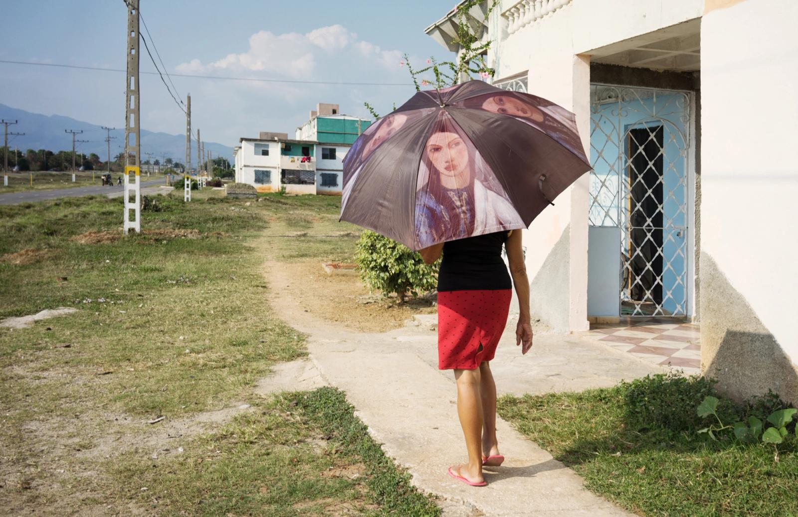 A woman walking down a path in a grassy field, wearing a red skirt and a black top, holding a black and white umbrella with a photo of a woman's face on it.