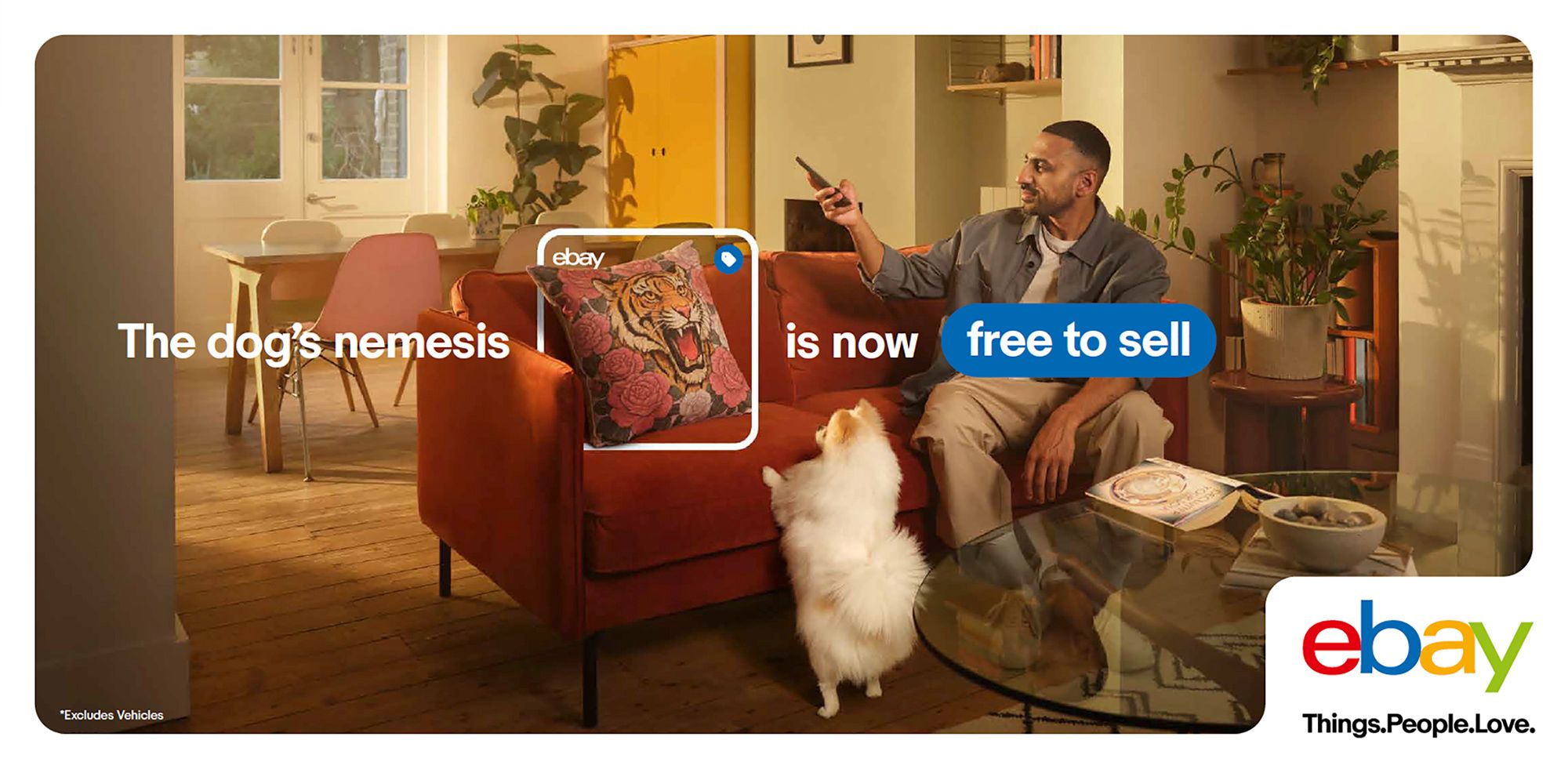A living room with a couch, a coffee table, and a man sitting on the couch with a phone in his hand, a dog sitting next to him.