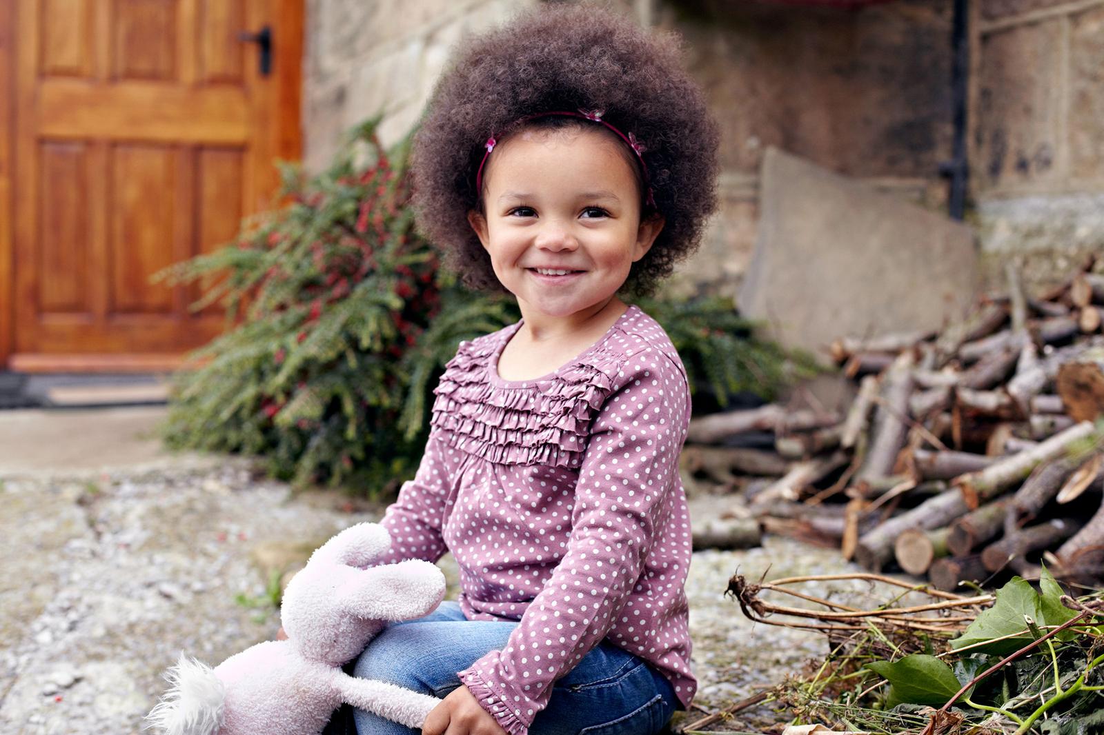 A young girl with curly brown hair and a big smile, wearing a pink sweater with white polka dots and blue jeans, sitting on the ground with a stuffed rabbit.