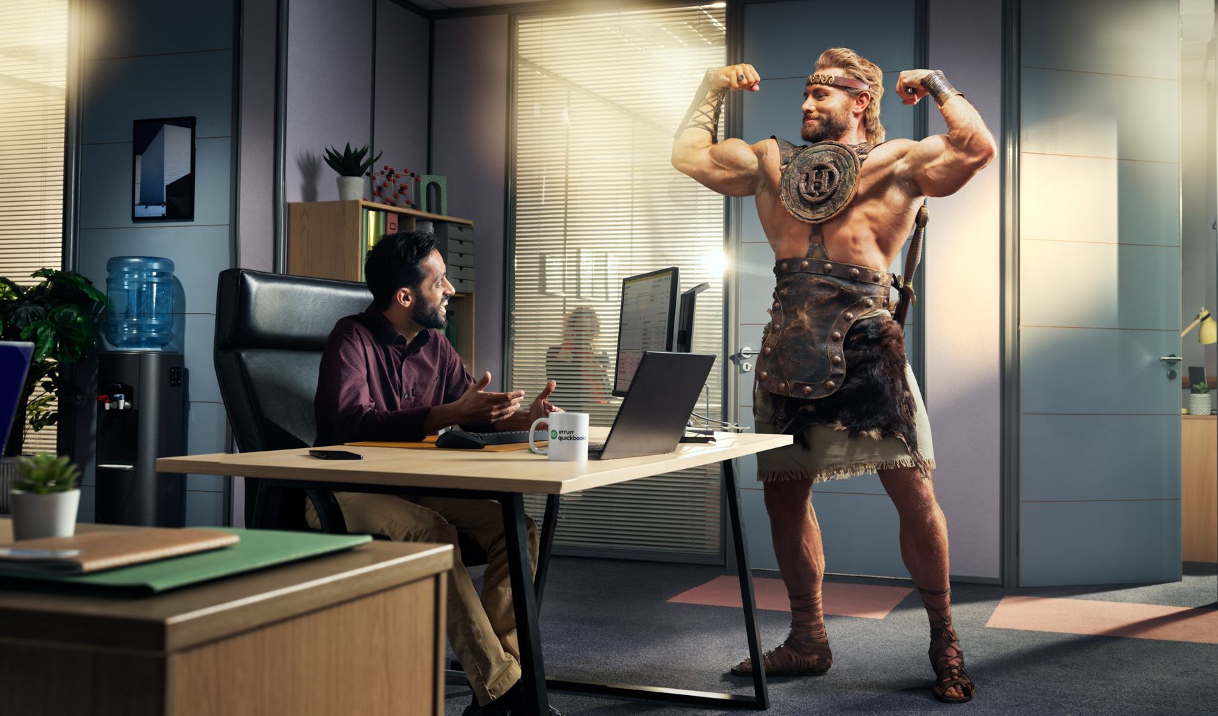 A man in a business office is interrupted by a muscular, ancient Greek warrior, dressed in a toga and carrying a sword, who is flexing his muscles and making a muscle.