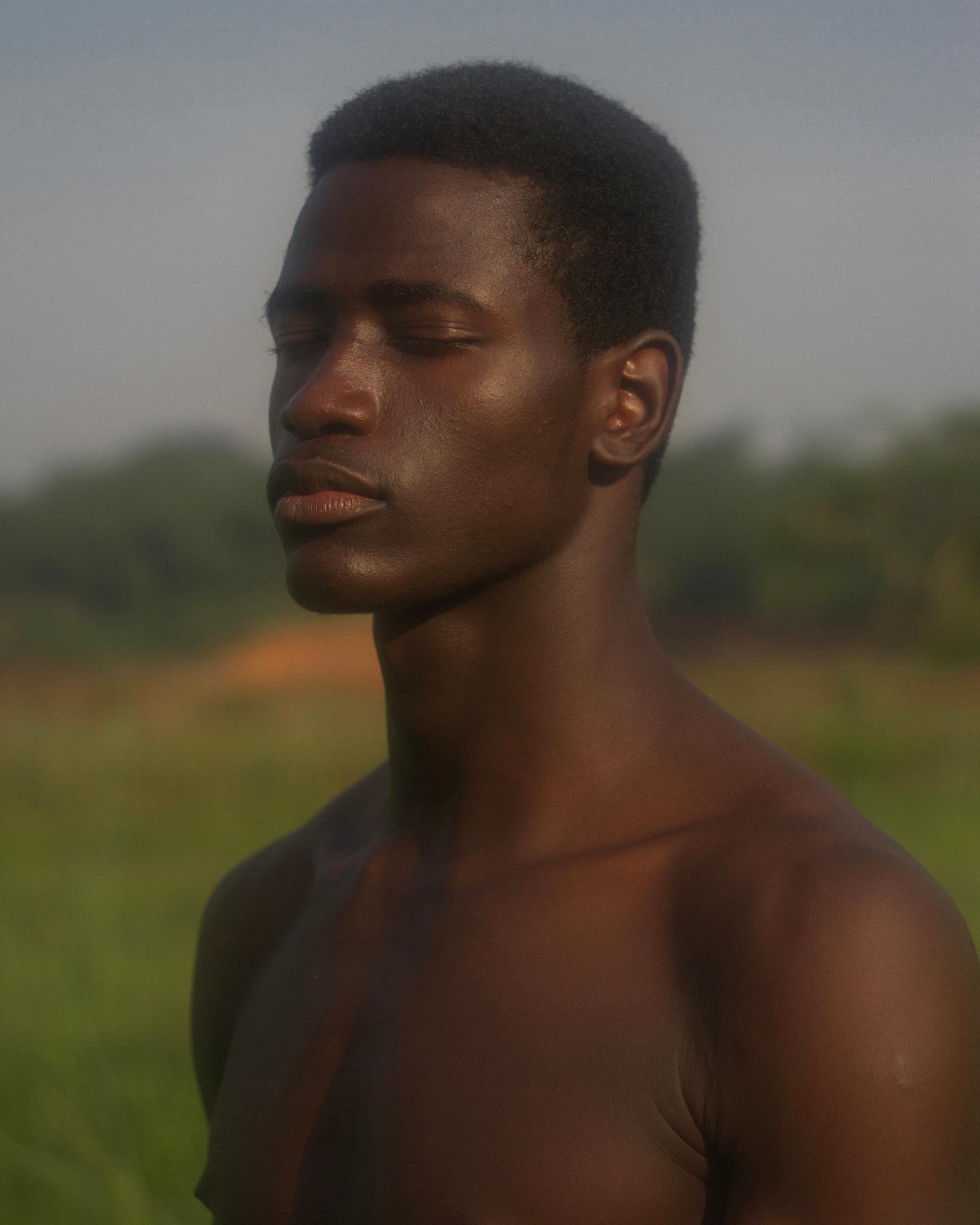 A young, shirtless, dark-skinned male with a short, dark, afro-like hair style, standing in a green, grassy field, with a blurred, green, forest-like background.
