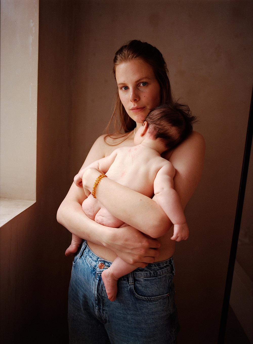 A young woman holding a naked infant in her arms, both of them standing in front of a plain, beige-colored wall.
