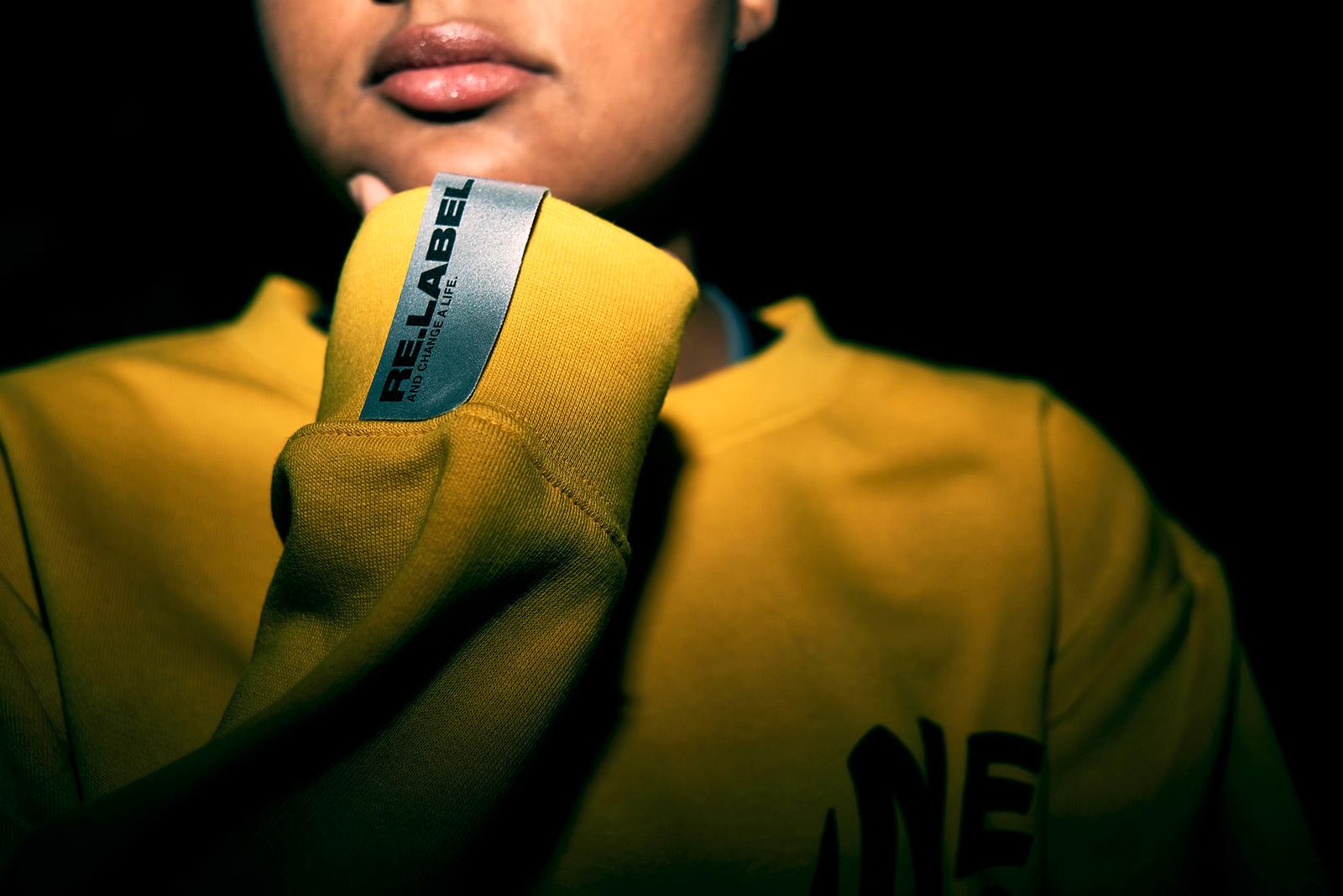 A woman wears a yellow sweatshirt with the label 'RE:LABEL AND CHANGE A LIFE.'.