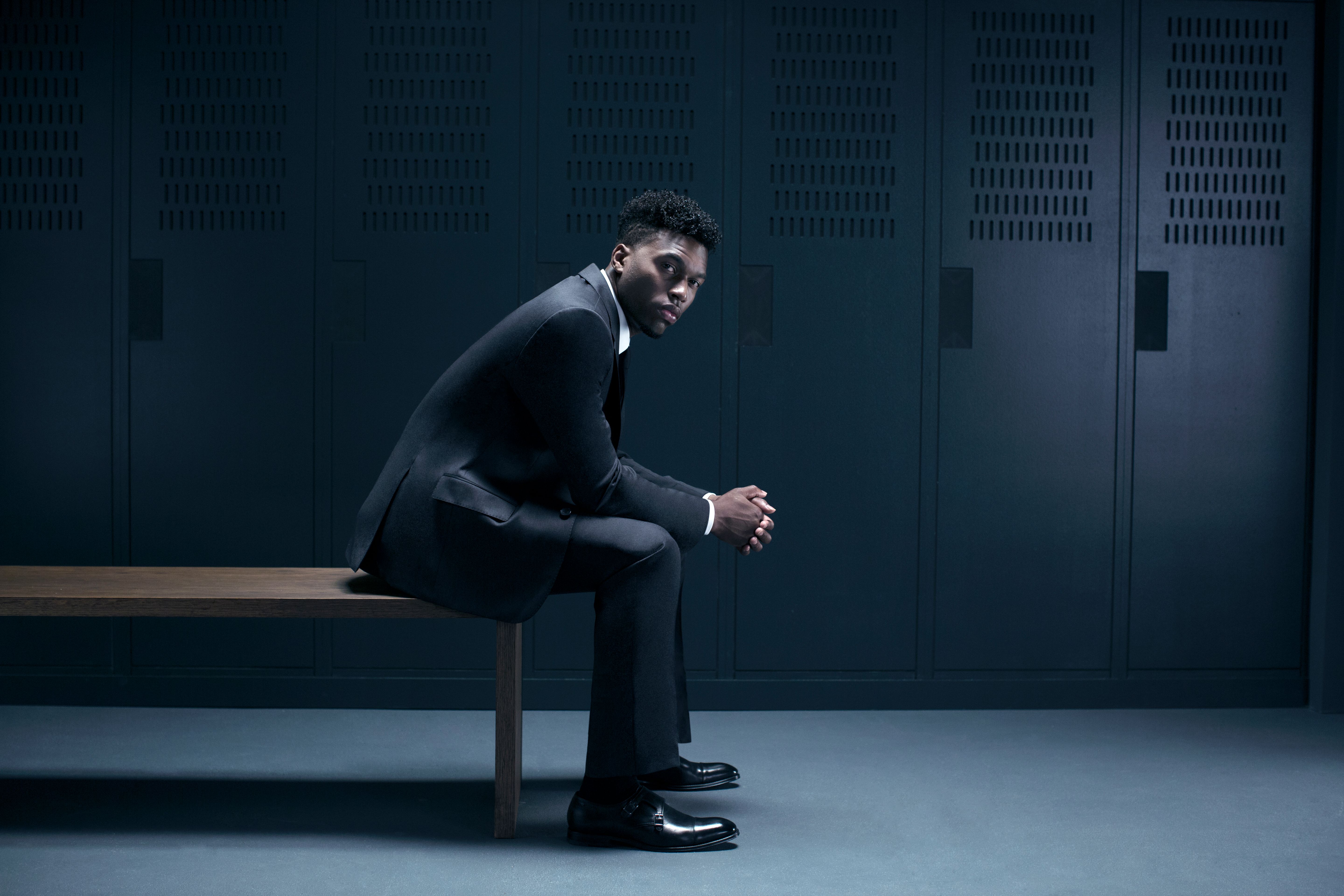 A young, African-American man in a suit and tie sits on a bench in a dimly lit, industrial-style locker room.