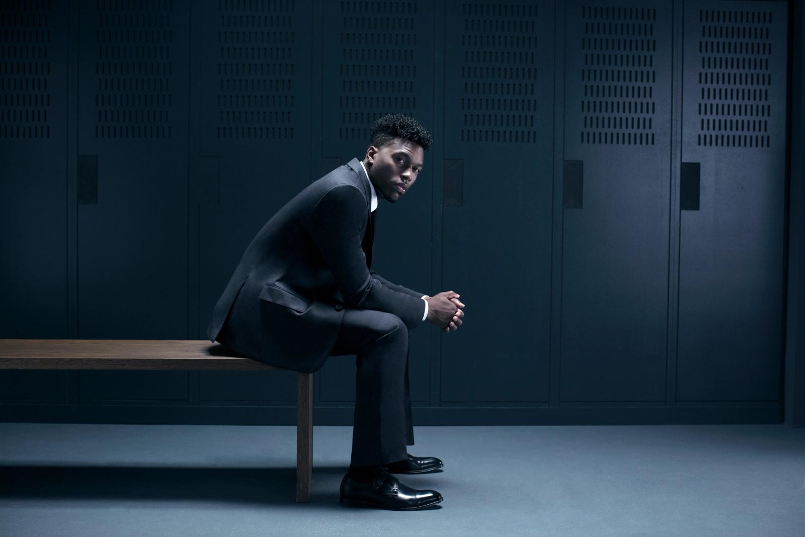 A young, African-American man in a suit and tie sits on a bench in a dimly lit, industrial-style locker room.