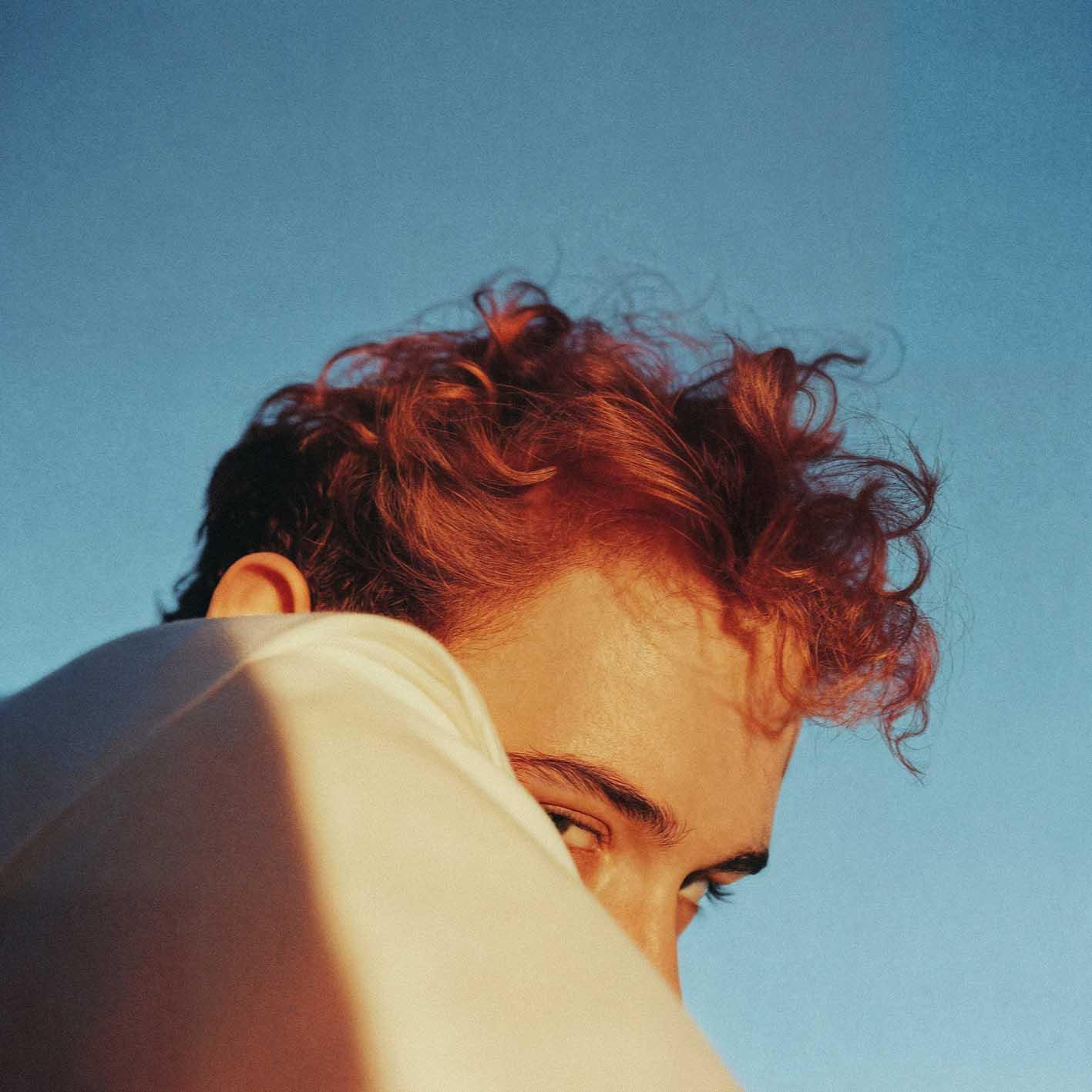 A person with short, messy red hair and a white shirt is looking over their shoulder, with the sun shining on their face.