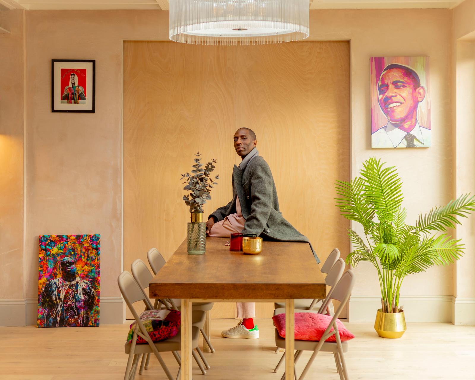 A man in a grey coat, pink trousers, and white sneakers sits at a wooden table, surrounded by artwork and plants, in a modern, light-wooden room.