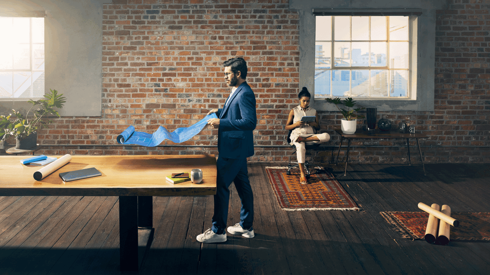 A man in a blue suit and white sneakers is holding a large blue architectural blueprint in a modern office with a woman sitting in the background, both in a professional setting.
