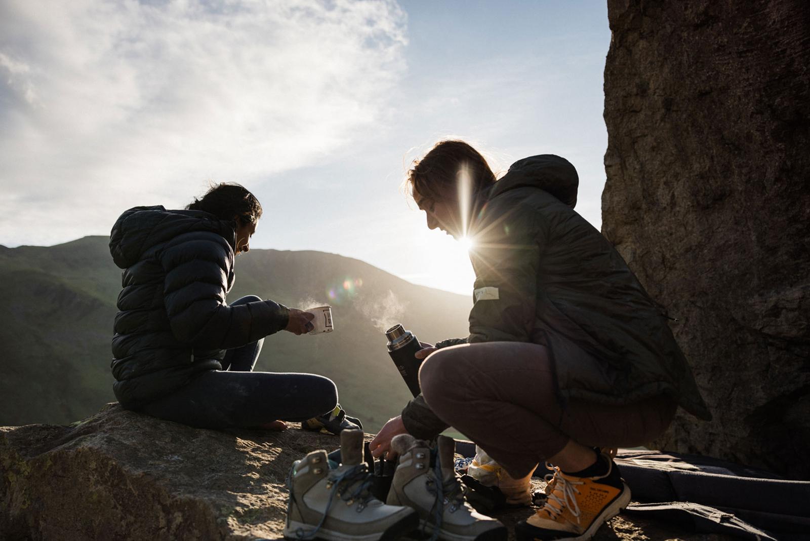 Two women in hiking boots and jackets sitting on a rock, looking out at a mountain landscape with a thermos and a cup of coffee in hand.