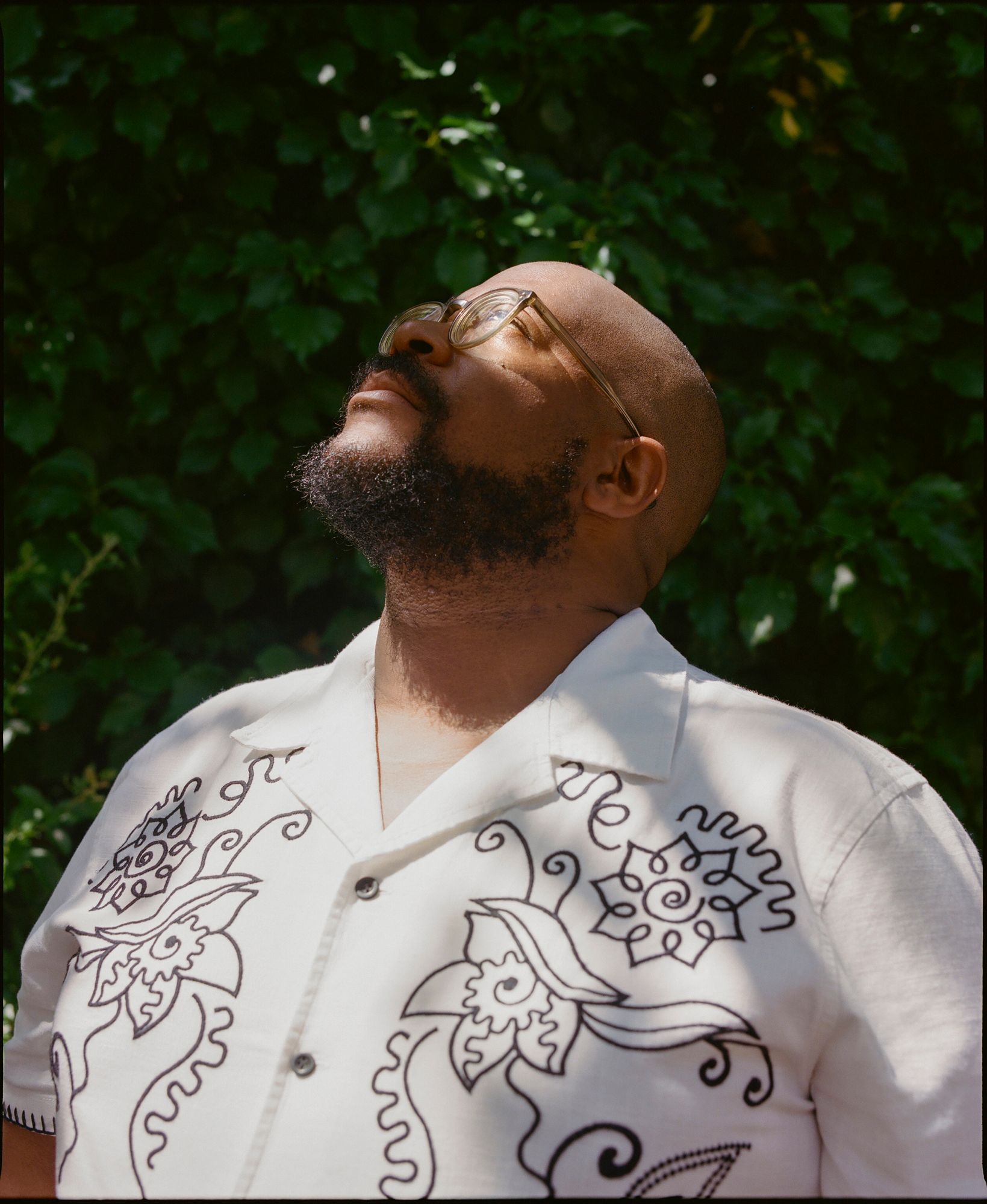 A man with a shaved head and a short, dark beard, wearing a white shirt with a black, intricate, hand-drawn design on the front, looking up towards the sky.