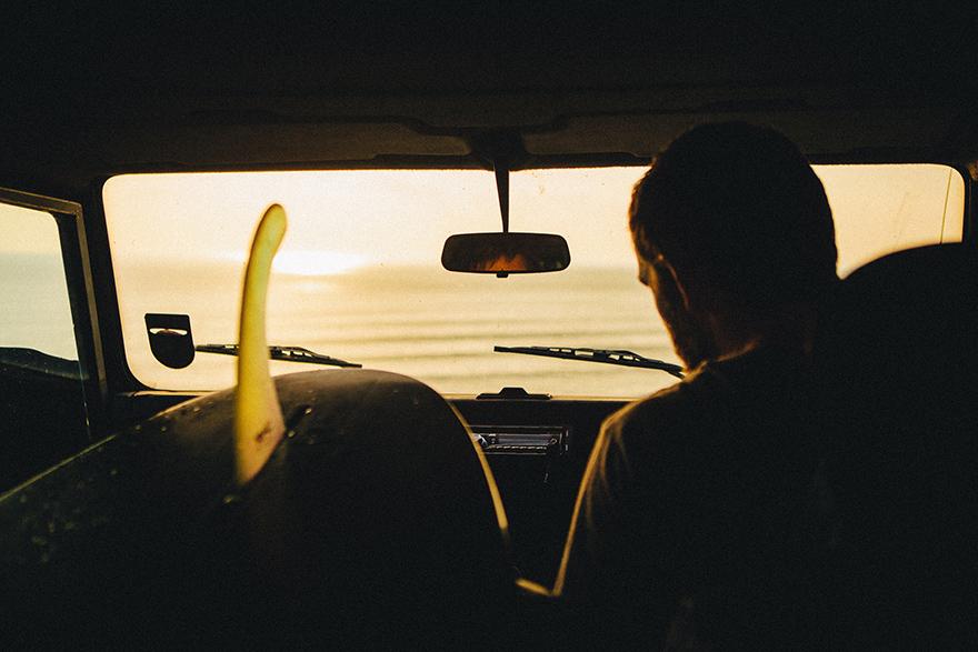 A person is sitting in the back of a car, looking out at the ocean through the rear window, with a surfboard visible on the roof.