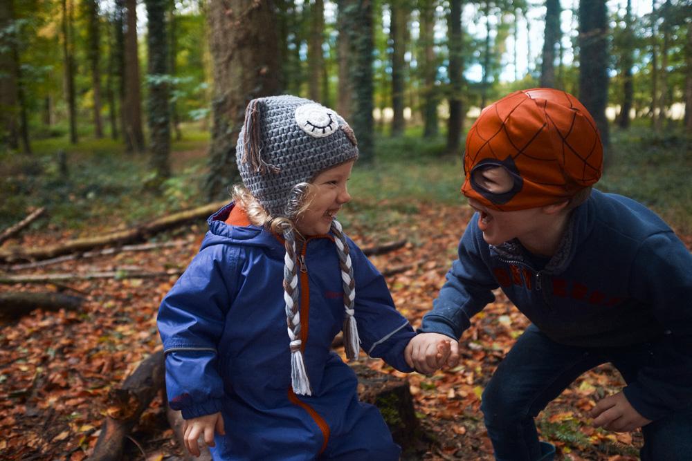 A young boy in a blue rain suit and a boy in a Spider-Man costume hold hands in a forest. The boys are smiling and looking at each other.
