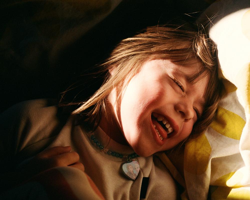 A young girl with a big smile and messy hair is lying on a bed, surrounded by pillows and a blanket, with a heart-shaped locket around her neck.