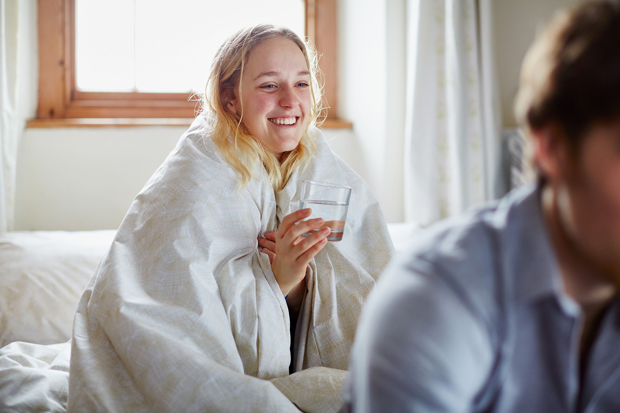 A woman with a messy, wet hair, wearing a white bathrobe, holding a glass of water, sitting on a bed, laughing at something in the background.