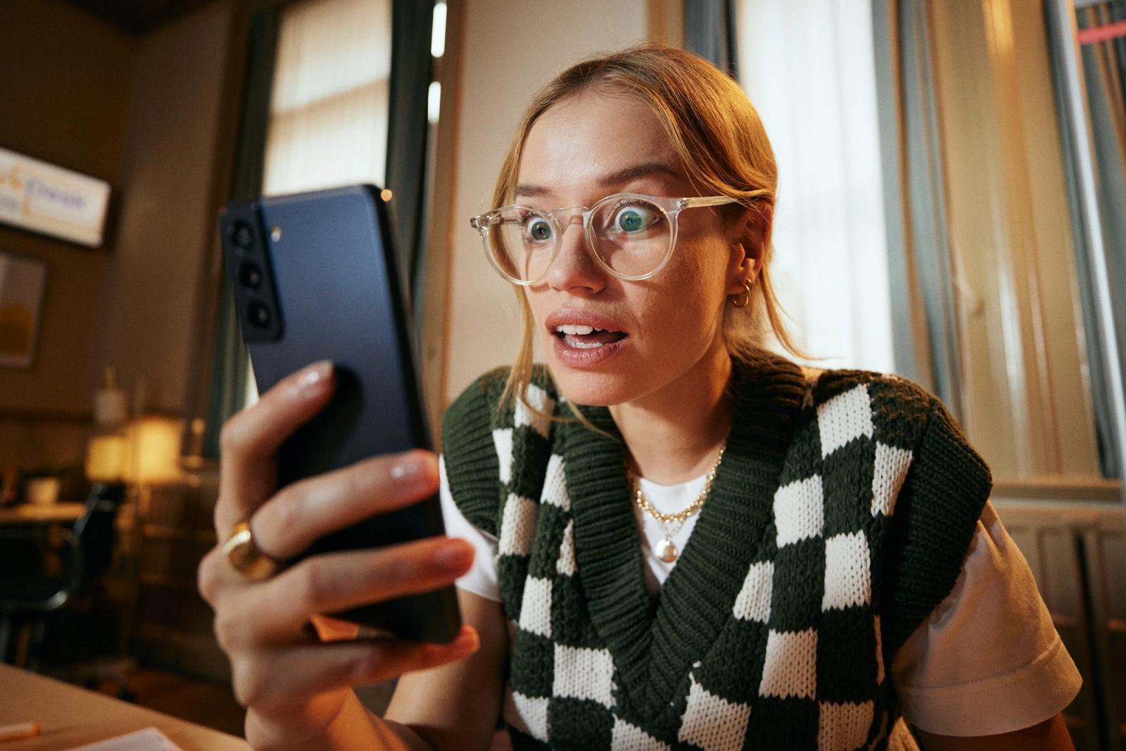 A woman with blonde hair and glasses is looking at her phone with a shocked expression. She is wearing a green and white argyle sweater and has a concerned look on her face.