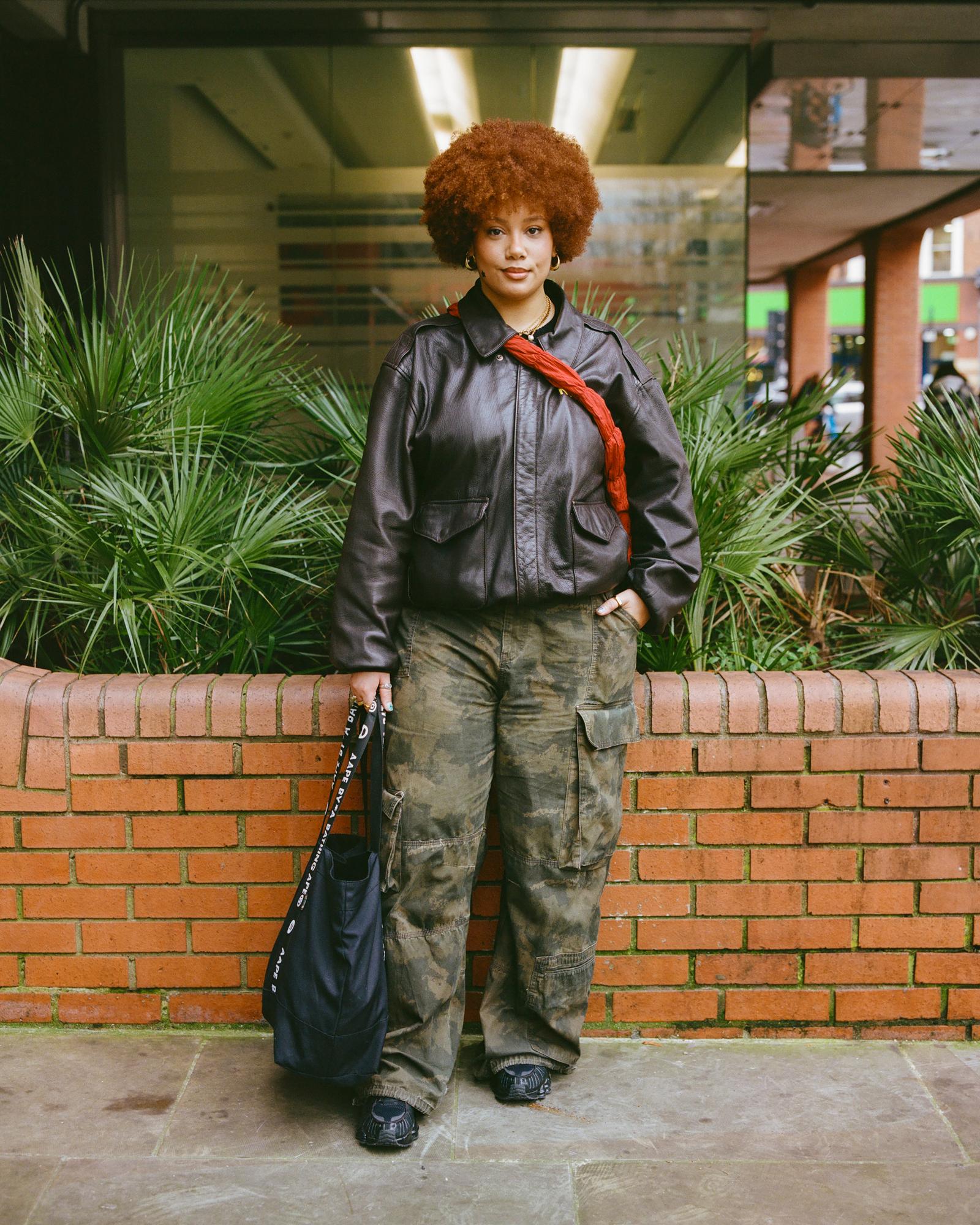 A woman with curly, red hair stands in front of a brick wall, wearing a black leather jacket, camouflage pants, and black boots, holding a black bag.
