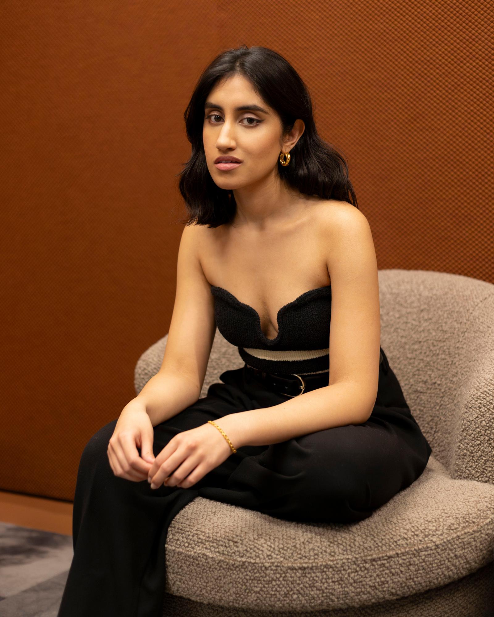 A woman with dark hair and a black dress sitting on a chair, looking directly at the camera.