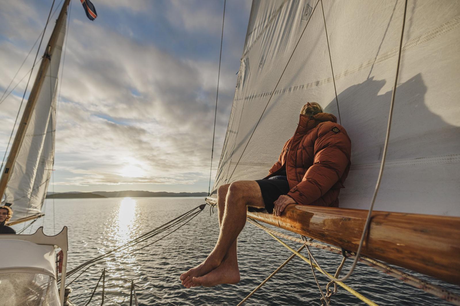 A man in a red jacket and black shorts is sitting on the bow of a sailboat, with the sun setting in the background.