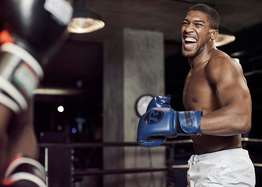 A shirtless, muscular black man in a boxing gym, wearing white shorts and blue boxing gloves, with a determined expression on his face.