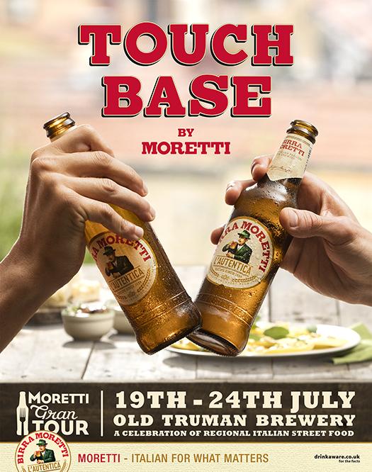 Two hands holding up bottles of Moretti beer, with a table of food in the background.