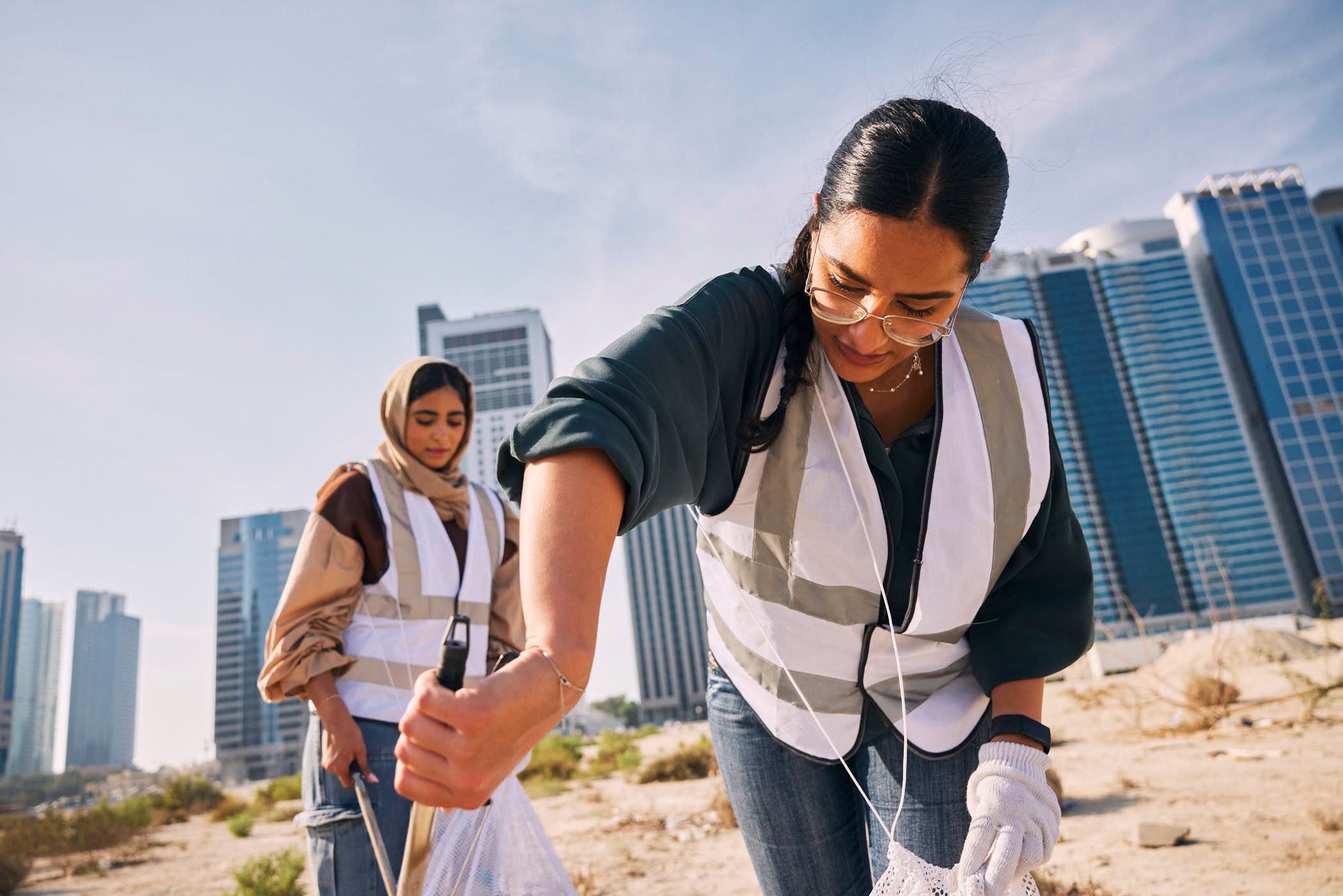 Two women in high-visibility vests, one wearing a hijab, stand in a dry, sandy area, picking up trash. They are both holding long-handled grabber tools and white trash bags, with.