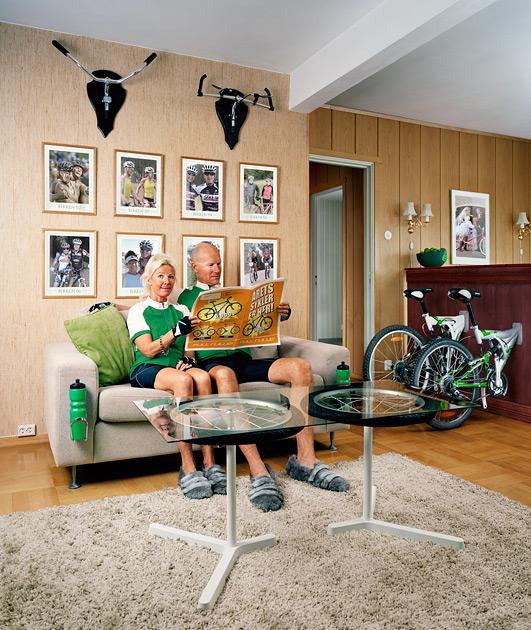 A couple in cycling gear sitting on a couch, reading a newspaper.