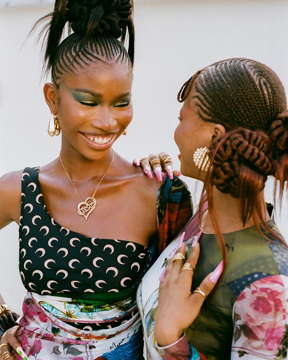 Two women with dark skin and long hair, both wearing gold jewelry and colorful clothing, smiling and embracing each other.