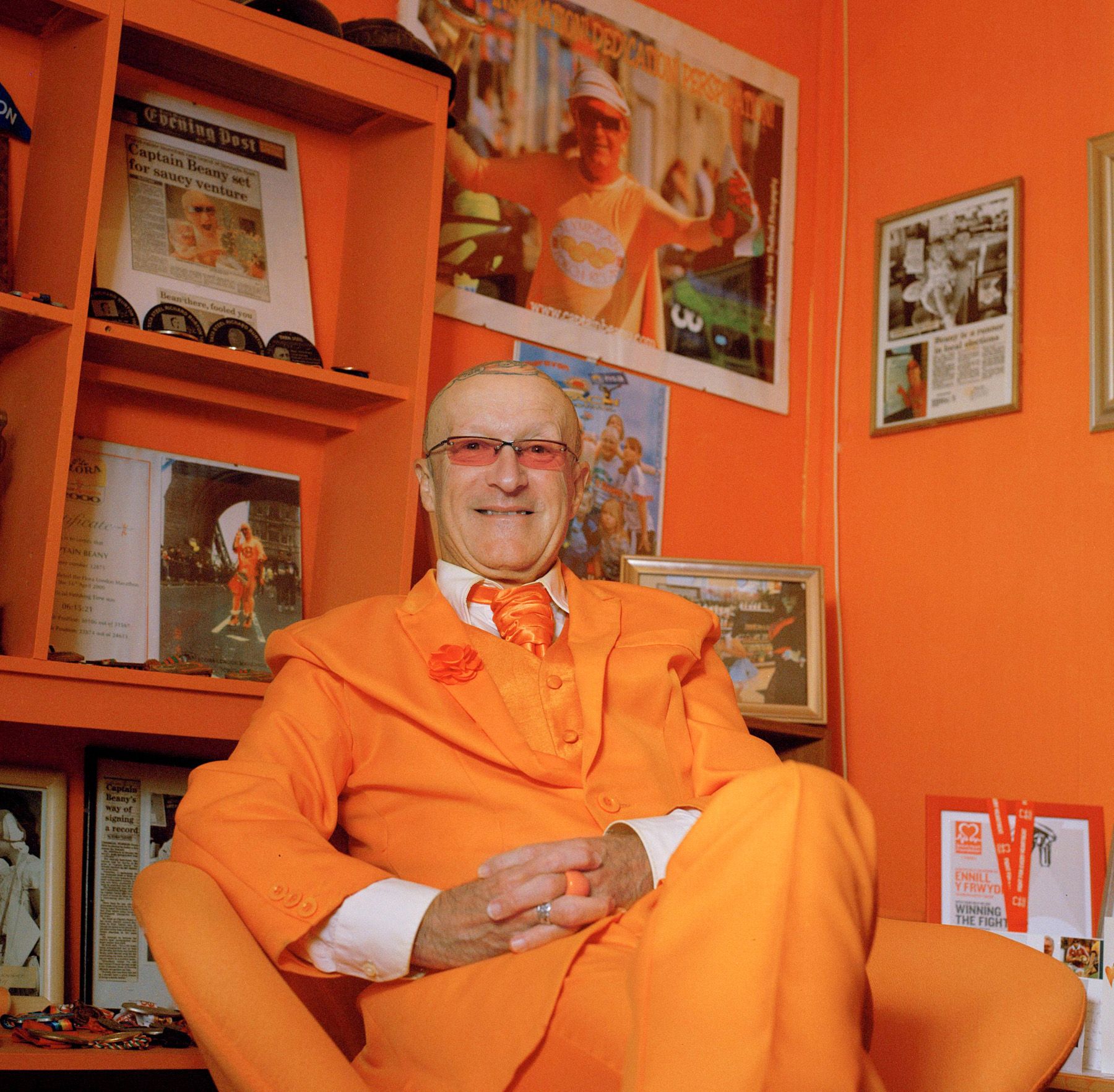 A man in a bright orange suit sits in a chair with objects and images on the walls and shelves behind him, including photographs and awards.