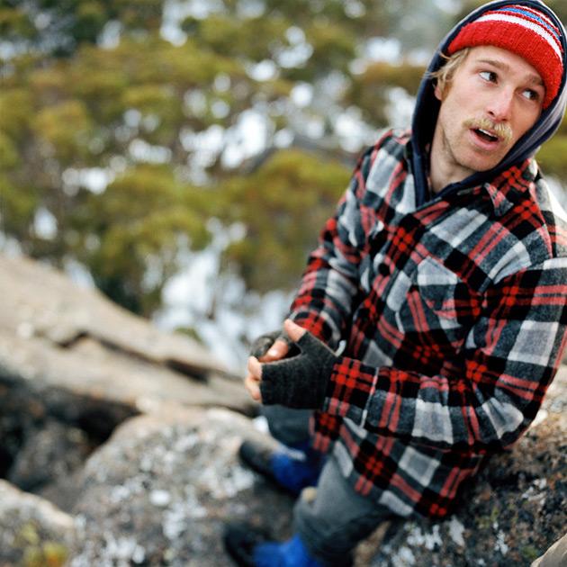 A man with a mustache and a red, white, and blue striped hat, wearing a red and black plaid flannel shirt, sitting on a rock in a forest.