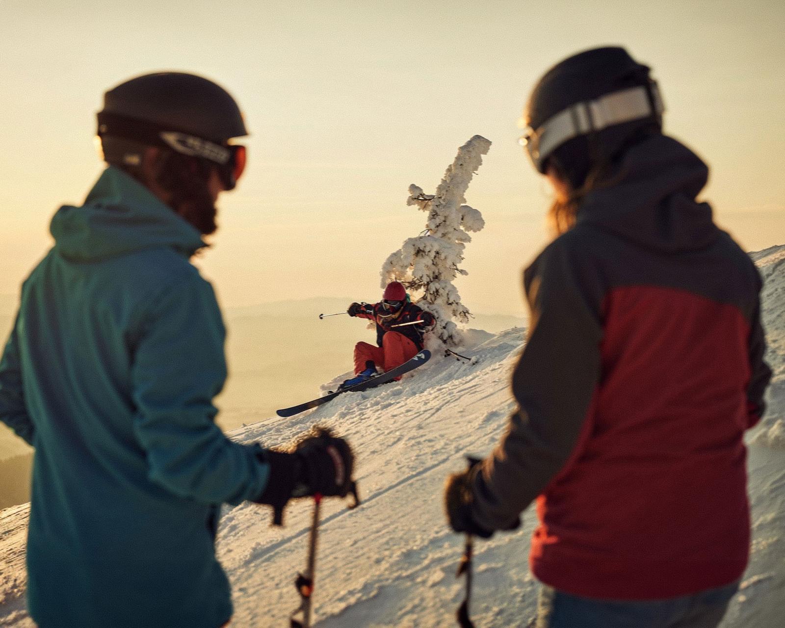 Two people in winter clothing and helmets are watching a skier in a red and black outfit sliding down a snowy mountain.