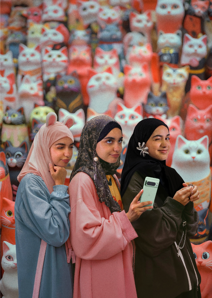 Three women in hijab and colorful clothing posing in front of a large collection of cat figurines and decorations.