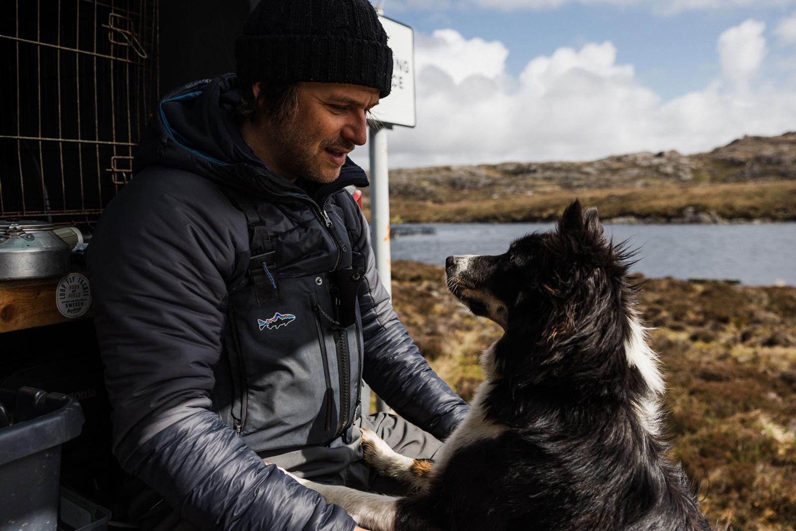 A man in a black beanie and a black and grey jacket is sitting on a rock, petting a black and white dog.