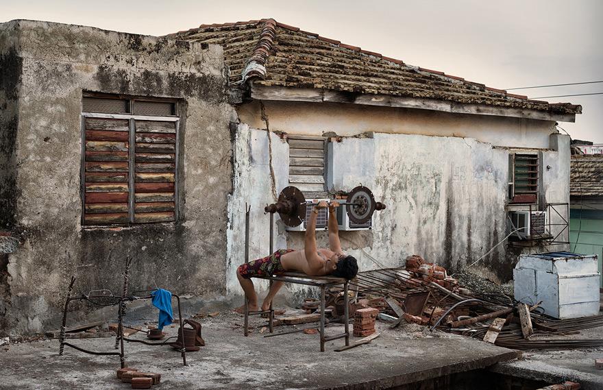 A man in a pair of floral shorts is lifting a weight on a makeshift outdoor gym in front of a dilapidated, old, abandoned building.