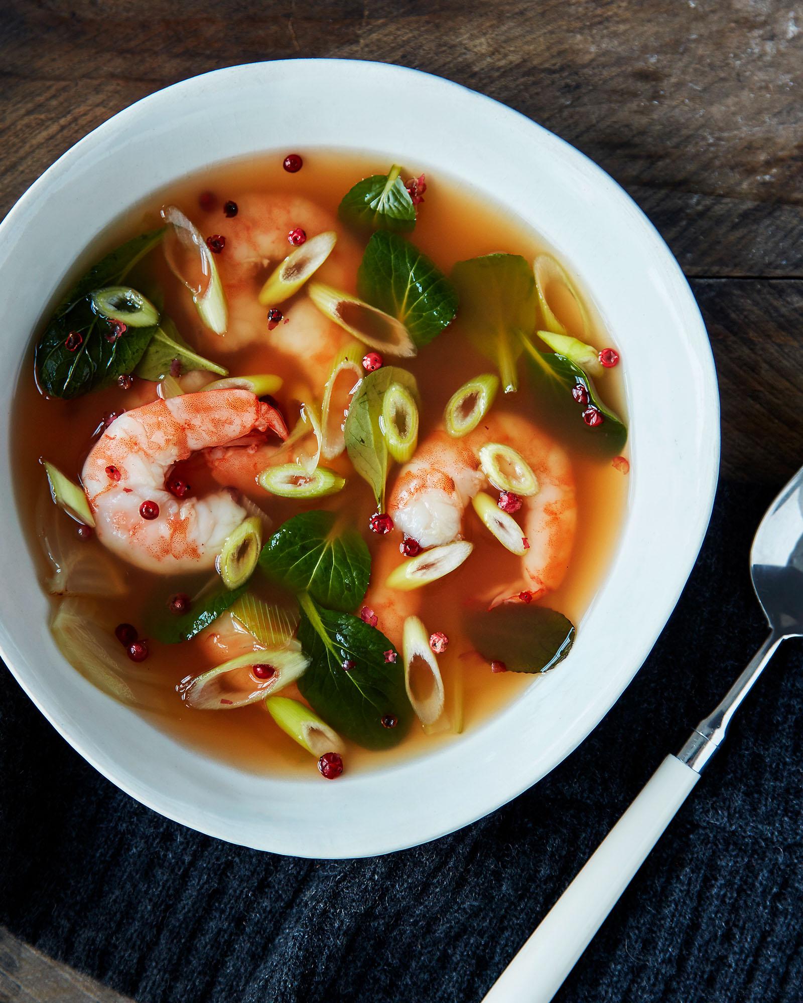 A bowl of soup containing prawns, vegetables, and a spoon.