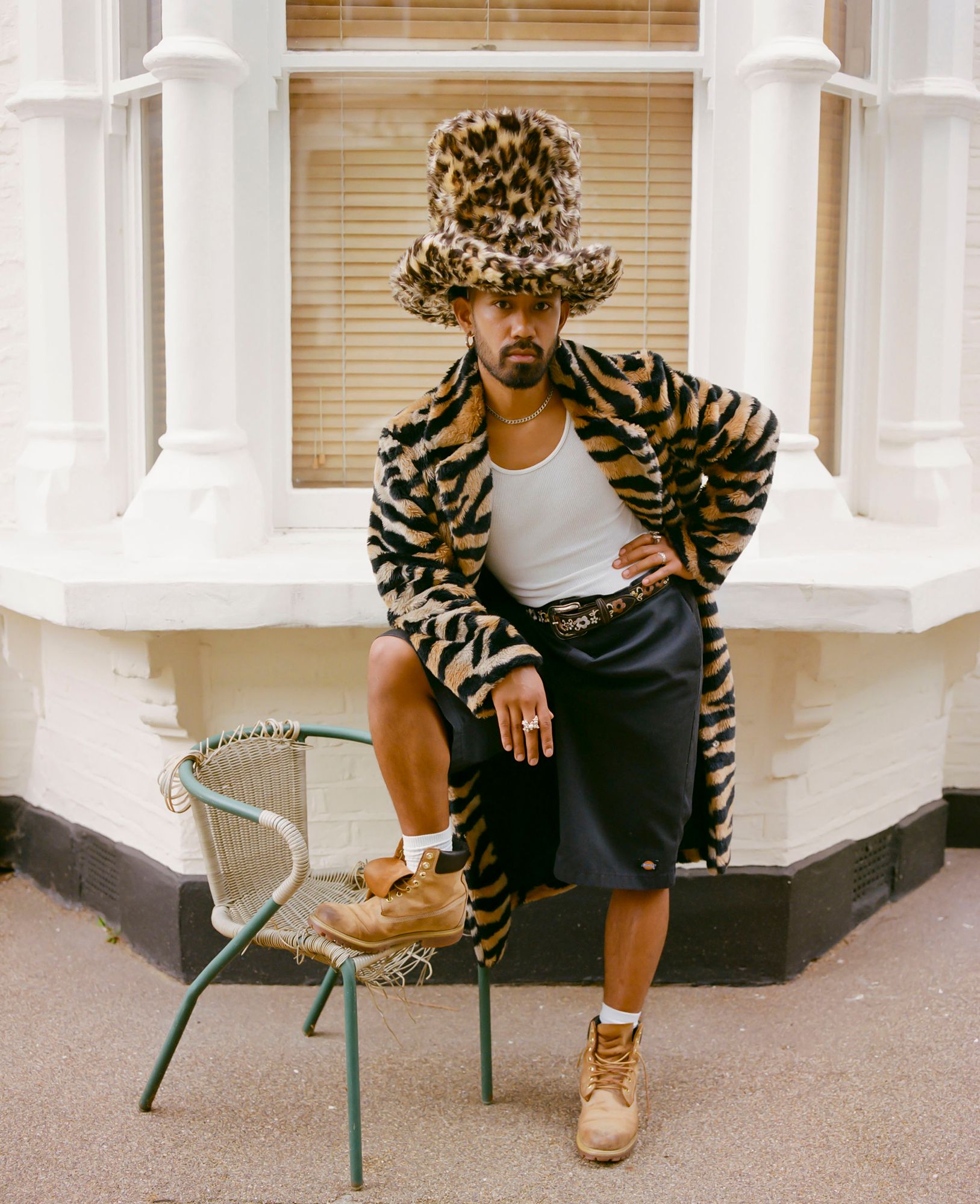 A man wearing a leopard-print coat, a white tank top, a black skirt, and a leopard-print hat, sitting on a chair in front of a white building with a window and a door.