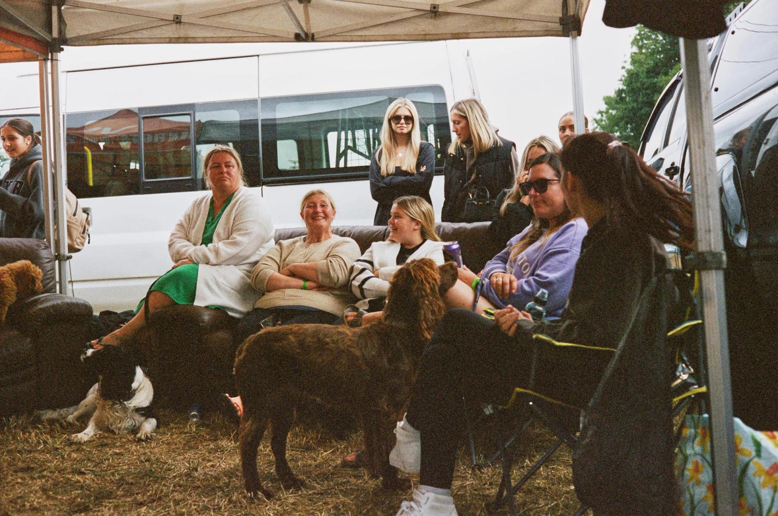 A group of women, some with dogs, sit under a tent with a white van and a bus in the background. They seem to be enjoying a gathering or event, possibly a festival or event, with.