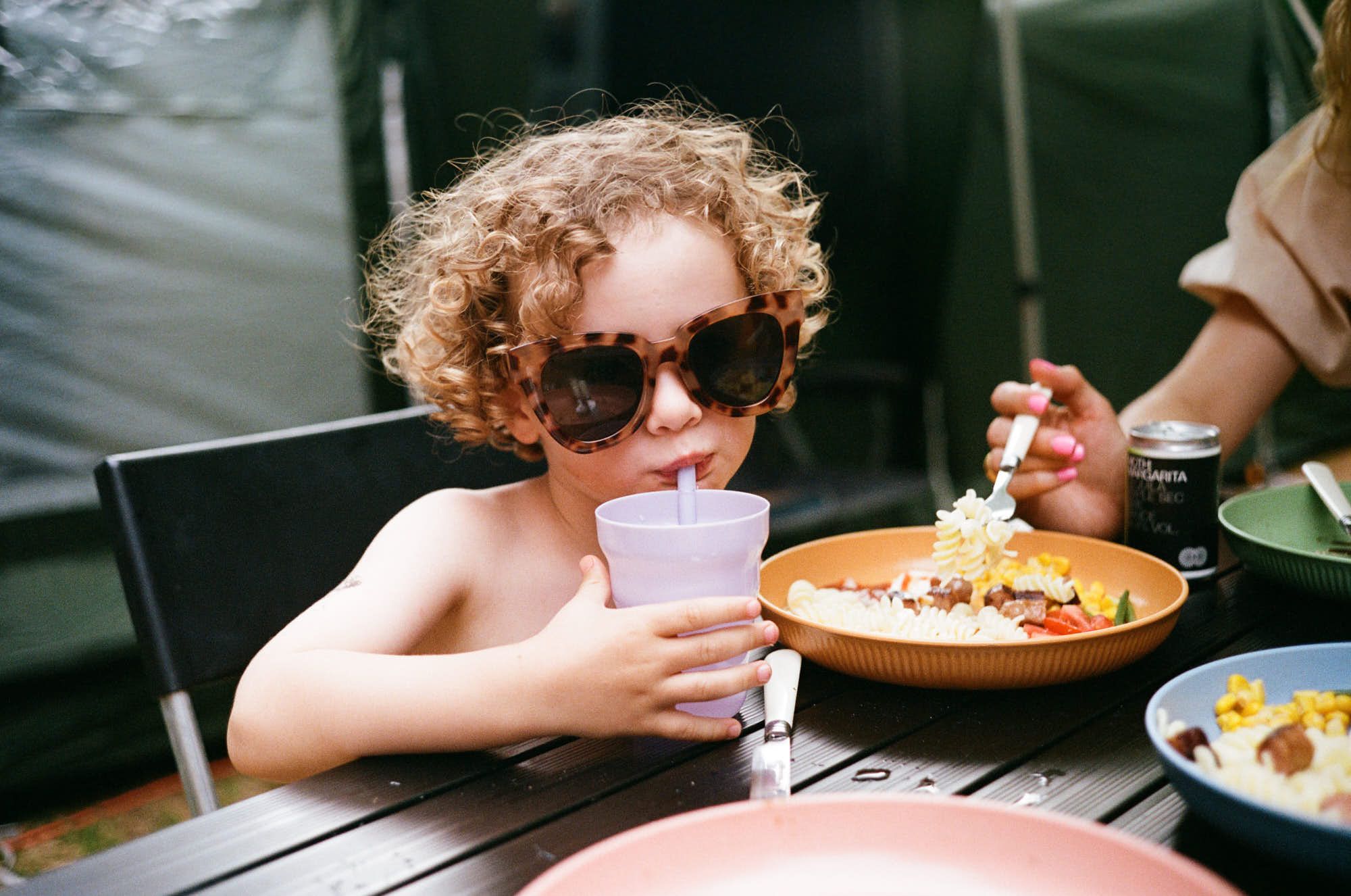 A young child with curly hair and sunglasses sits at a table, drinking from a straw while eating a meal. The child's meal appears to be a bowl of pasta with vegetables.