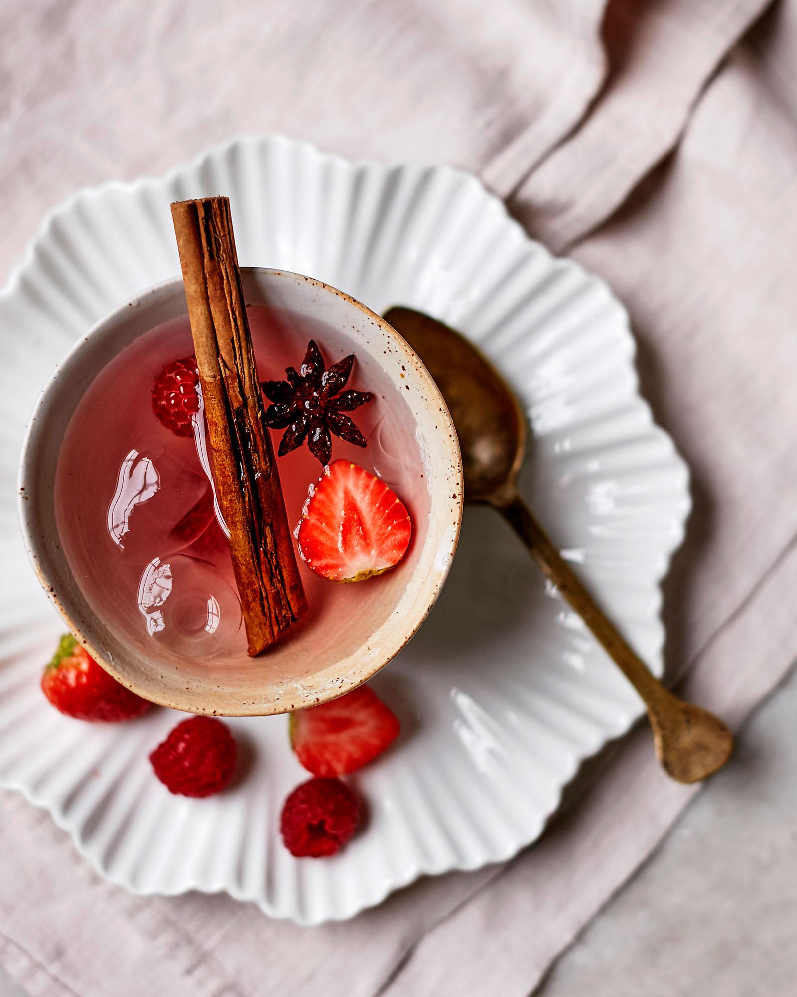 A ceramic cup with a pink spiced berry drink, garnished with a cinnamon stick, strawberry, and raspberries, on a white scalloped plate.
