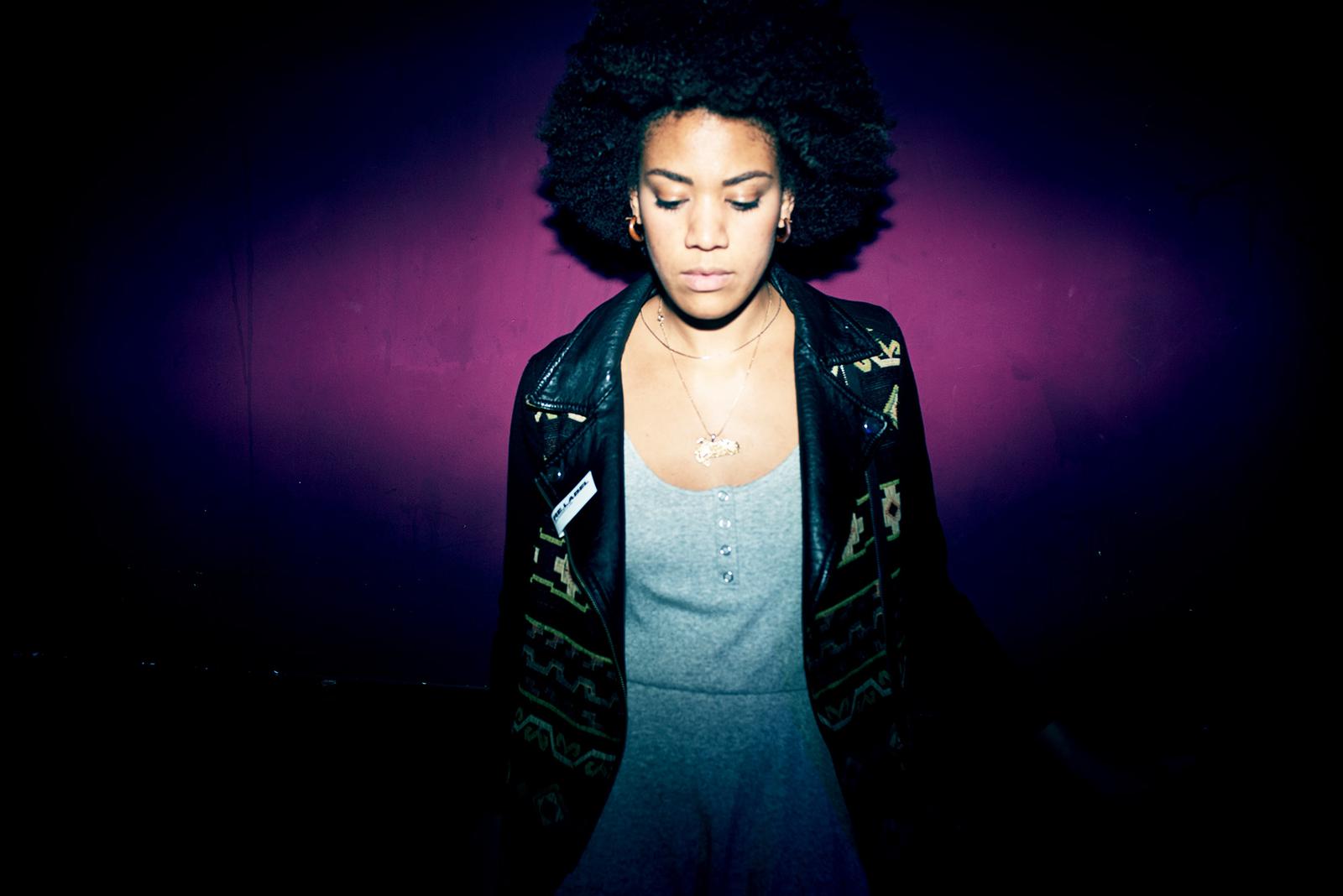 A woman with curly, dark hair and a black leather jacket, wearing a grey dress and a silver necklace.