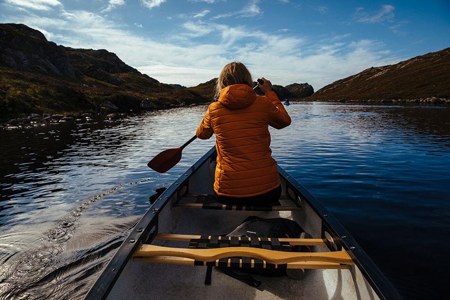 A woman in an orange jacket paddles a canoe through a serene lake surrounded by mountains.